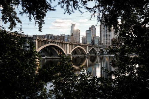 A bridge arches over a river, framed by trees, with a city skyline in the background under a clear sky.