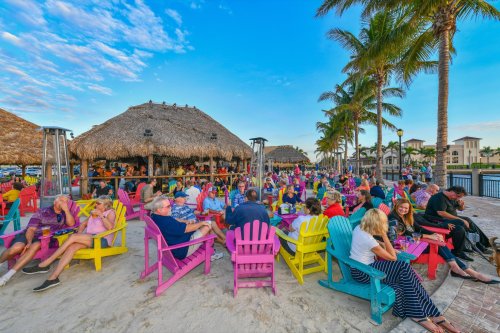 A vibrant beach scene with colorful chairs, people socializing, and palm trees under a clear blue sky.