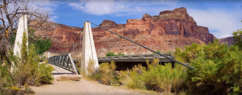 A suspension bridge spans a dry riverbed, framed by red rock cliffs and green vegetation under a blue sky.