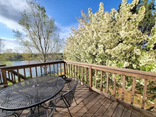 A wooden deck with a round table and chairs overlooks a lake and blooming trees under a clear blue sky.