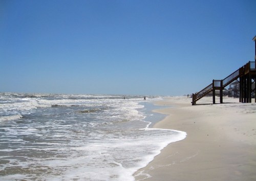 A sandy beach with gentle waves, a clear blue sky, and a wooden staircase leading to a beach house in the background.