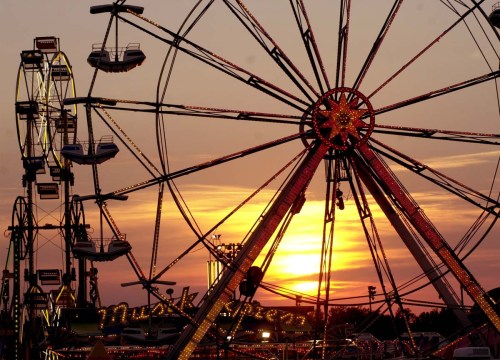A vibrant sunset behind a Ferris wheel at a carnival, with colorful lights illuminating the scene.