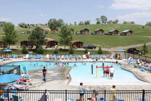 A busy outdoor pool area with people swimming, surrounded by lounge chairs and cabins on a grassy hill in the background.