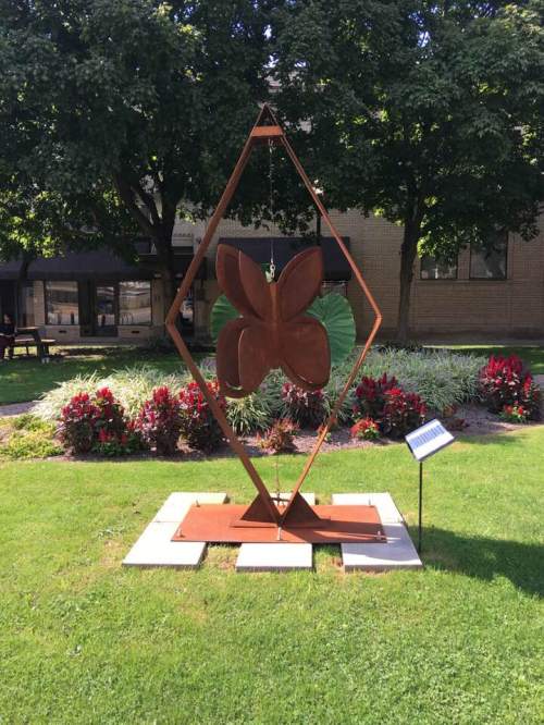A large, rust-colored butterfly sculpture stands in a green park surrounded by flowers and trees.