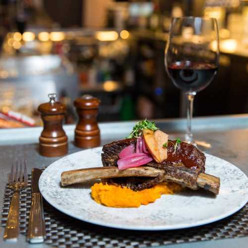 A gourmet plate featuring a ribeye steak, sweet potato puree, pickled vegetables, and a glass of red wine.