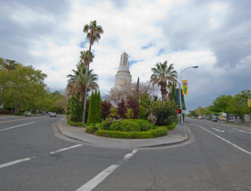 A roundabout with lush greenery and palm trees, featuring a tall tower in the background under a cloudy sky.