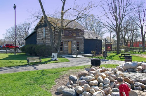 A rustic log cabin with a porch, surrounded by trees and a grassy area, with a child playing near a rock formation.