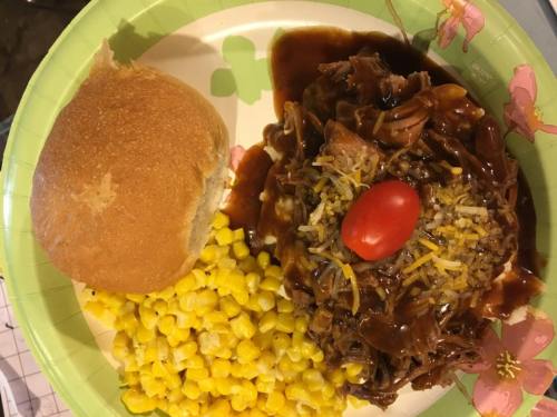 A plate with shredded meat topped with cheese and a cherry tomato, served with corn and a dinner roll.