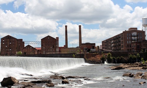 A scenic view of a waterfall in front of old industrial buildings under a partly cloudy sky.