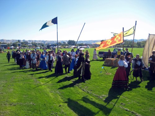 A group of people in historical costumes parades on a grassy field, with flags and tents in the background.
