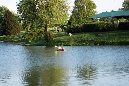 A person kayaking on a calm lake, with trees and a park shelter in the background.