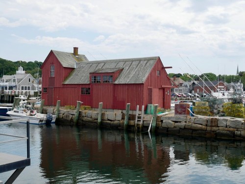 A red wooden building by the water, surrounded by boats and a scenic harbor with distant houses and trees.