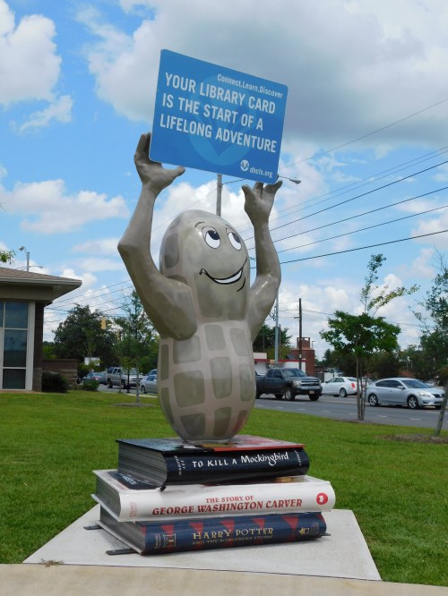 A cheerful peanut statue holds a sign about library cards, standing on stacked books in a grassy area.