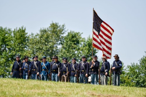 A group of Civil War reenactors in uniform, holding an American flag, standing on a grassy hill with trees in the background.