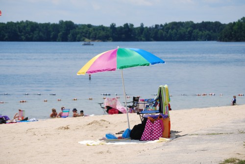 A colorful beach umbrella shades a chair on the sandy shore, with people enjoying the water in the background.