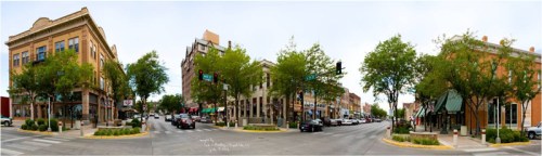 A panoramic view of a bustling city intersection with shops, trees, and cars under a cloudy sky.