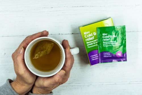 A person's hands holding a cup of tea next to a box and packet of cold and flu relief medication.