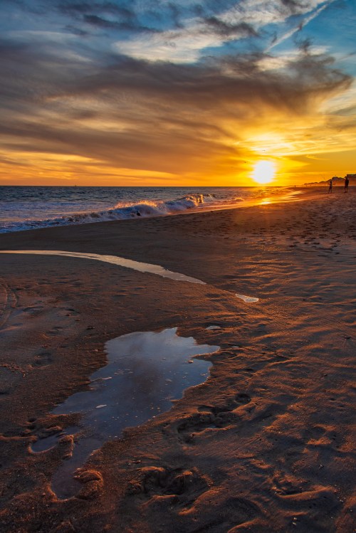 Sunset over a beach with gentle waves, a puddle reflecting the sky, and silhouettes of people walking along the shore.