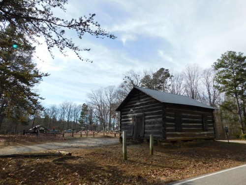A rustic log cabin surrounded by bare trees and a gravel path, under a cloudy sky.