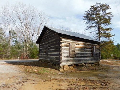 A rustic log cabin with a metal roof, set near a road and surrounded by bare trees and fallen leaves.