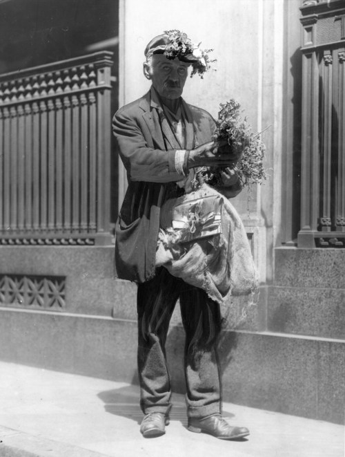 A man in vintage clothing stands on a street, holding a bundle of plants and wearing a floral headpiece.