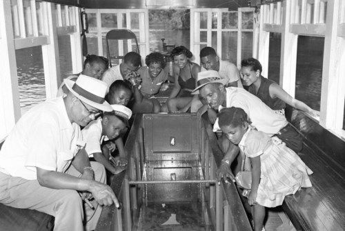 A group of people, including children, gathered on a boat, looking at something in the center, with water in the background.