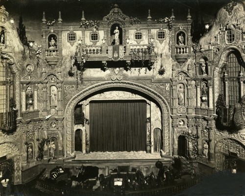 A vintage theater interior featuring an ornate stage with intricate decorations and a closed curtain.