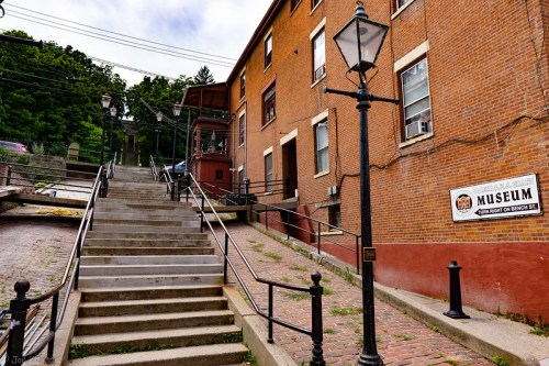 Stairs leading to a brick building with a museum sign, surrounded by greenery and vintage street lamps.