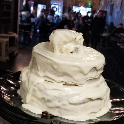 A three-tiered cake with white icing, slightly melting, displayed on a black plate in a busy restaurant setting.