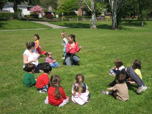 A group of children sitting on grass, with adults engaging them in a playful activity in a sunny park.