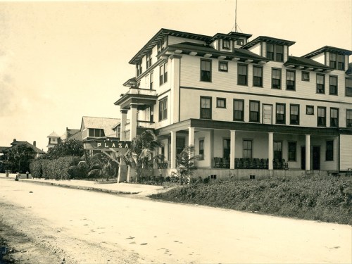 Historic photo of the Plaza Hotel, featuring a three-story building with a sign and palm trees along a dirt road.