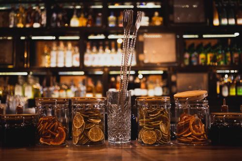 A bar countertop with jars of dried citrus slices and cocktail tools, set against a blurred backdrop of bottles.