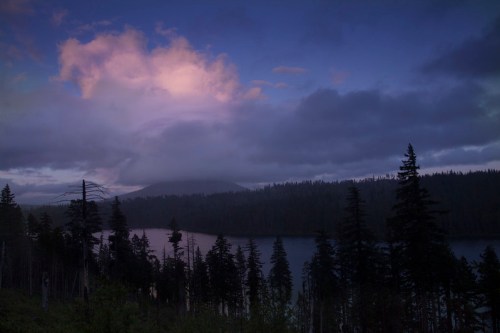 A serene lake surrounded by tall trees, with a misty mountain and colorful clouds in the twilight sky.