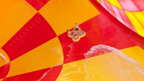 A group of people on a yellow inflatable raft sliding down a colorful water slide with splashing water.