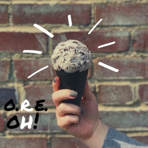 A hand holds a cone of cookies and cream ice cream against a brick wall, with playful text saying "O.R.E. OH!"