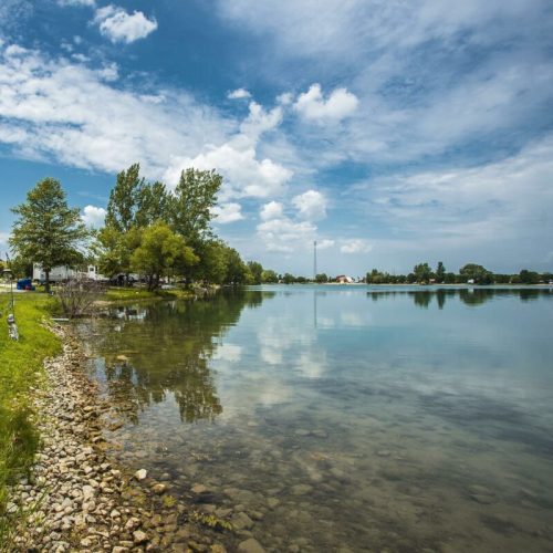 A serene lakeside scene with trees, a clear sky, and reflections on the water's surface.