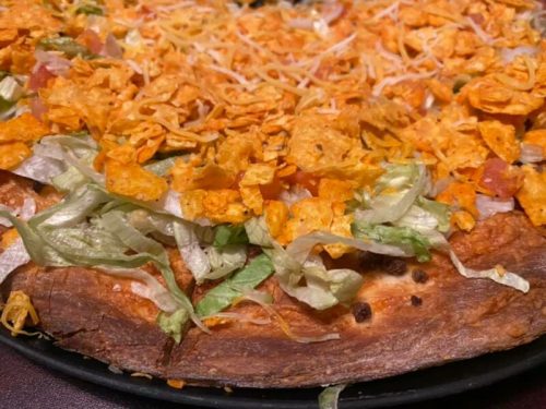Close-up of a pizza topped with lettuce, tomatoes, cheese, and crushed tortilla chips on a golden crust.