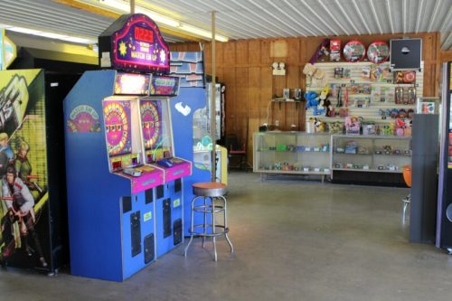 A colorful arcade game machine in a rustic game room with shelves of toys and collectibles in the background.
