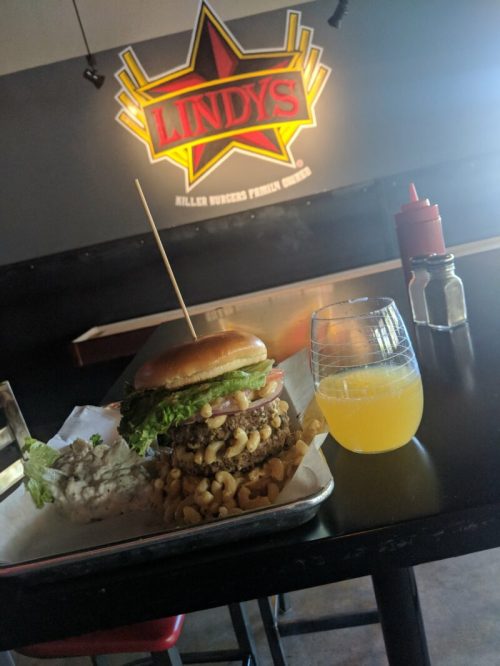 A towering burger with lettuce and a side of macaroni, accompanied by a glass of orange drink, set in a restaurant.