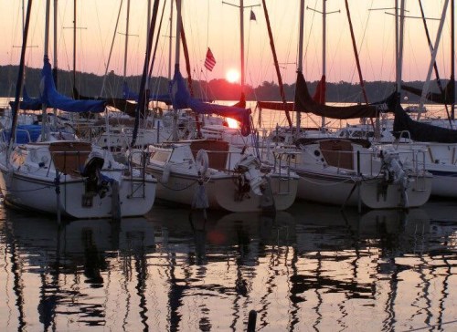 Sailboats docked at sunset, with reflections on the water and a warm orange sky in the background.