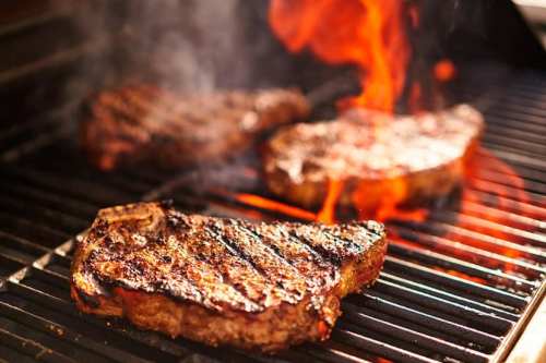 Grilled steaks sizzling on a barbecue, with flames and smoke rising around them.
