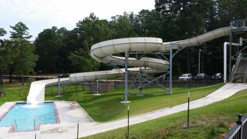 A water slide spirals down into a small pool, surrounded by green grass and trees in a recreational area.