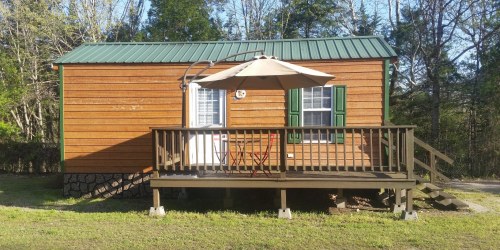 A small wooden cabin with a green metal roof, featuring a deck, umbrella, and two chairs in a natural setting.