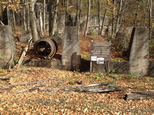 Abandoned site with a rusted barrel, concrete remnants, and a sign among fallen leaves and trees in a forested area.