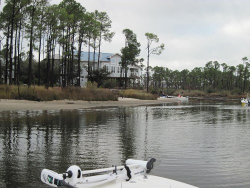 A boat on calm water near a sandy shore, with tall trees and a house in the background under a cloudy sky.
