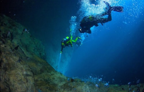 Two divers explore an underwater cave, surrounded by bubbles and illuminated by their flashlights.