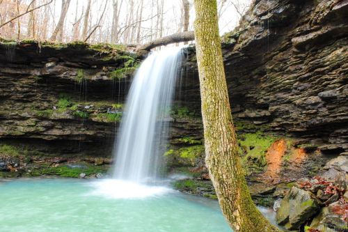 A serene waterfall cascades over rocky cliffs into a turquoise pool, surrounded by mossy stones and trees.