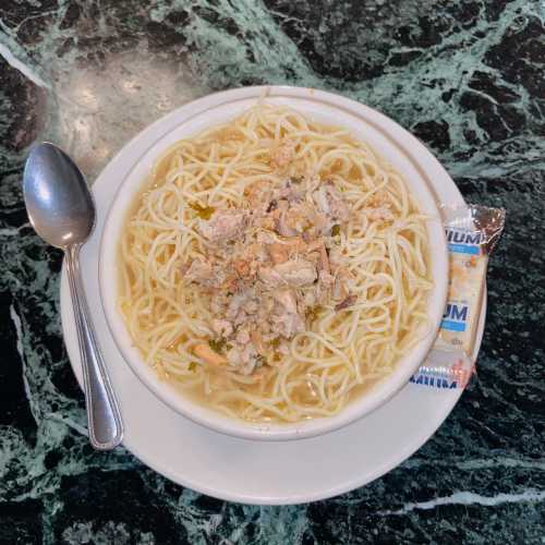 A bowl of noodles topped with shredded meat, served on a marble table with a spoon and a packet of seasoning.