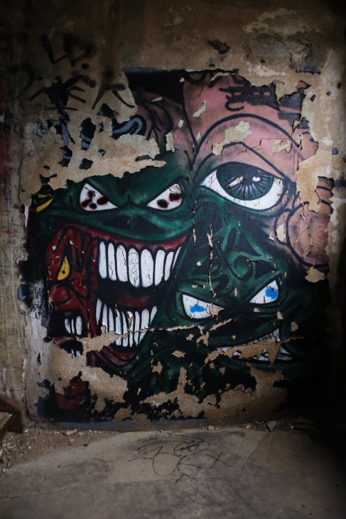 A vibrant graffiti mural featuring distorted faces with exaggerated features and multiple eyes on a weathered wall.