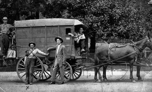 Two men in suits stand beside a horse-drawn wagon, with two others seated inside and on top, in a vintage setting.
